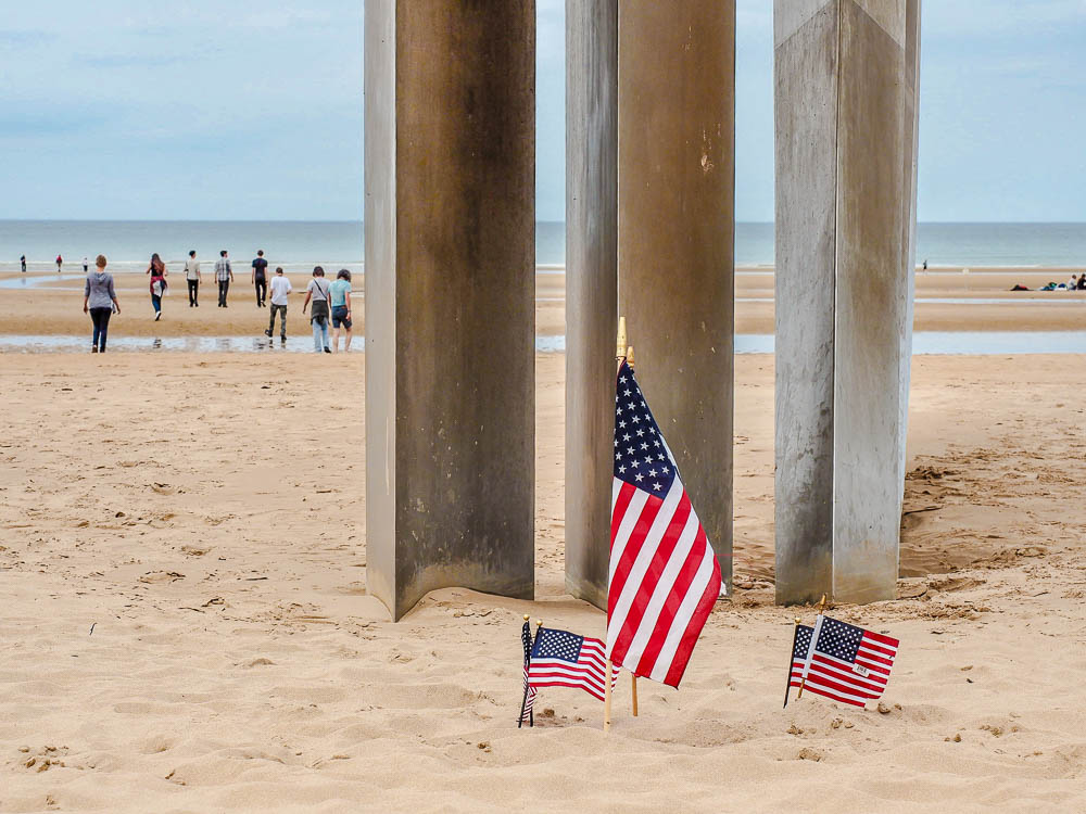 three small american flags in front of a memorial on omaha beach with a group of teenagers in the background