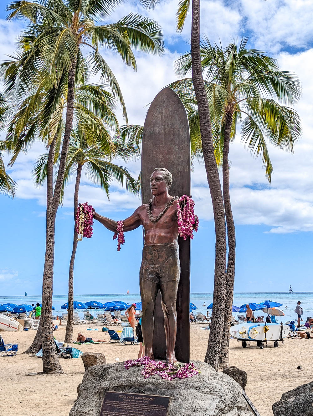 statue of a surfer holding pink flower leis on a beach